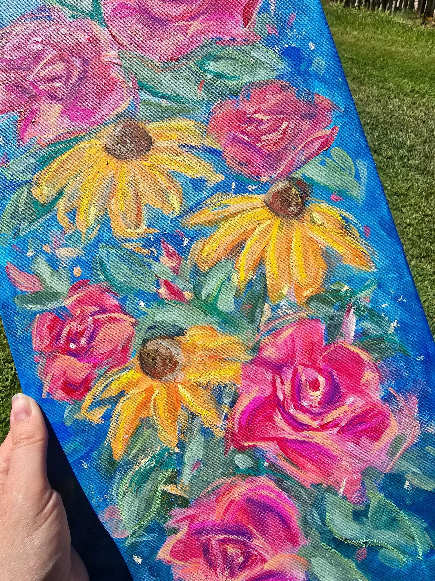 A hand holding a rectangular canvas painting outdoors over green grass. The painting features bright pink roses and yellow coneflowers with green leaves on a vivid blue background.