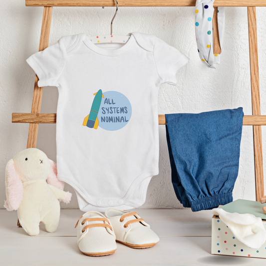 Baby onesie with 'All Systems Nominal' and accessories including pants, shoes, and a plush toy on a wooden stand.