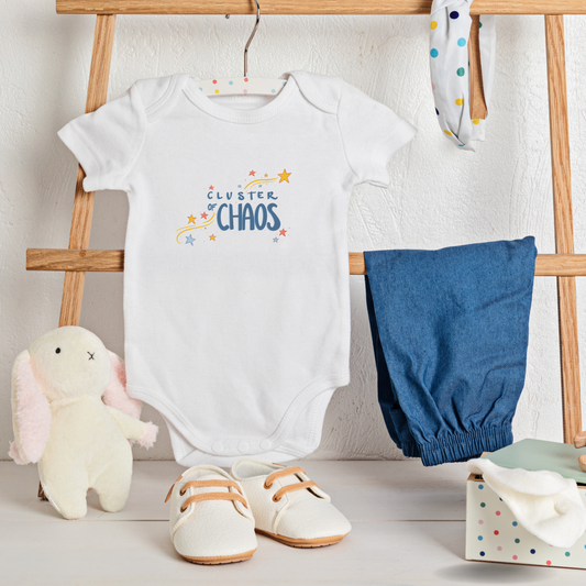 Baby onesie with text 'Cluster of Chaos' and starts and accessories including pants, shoes, and a plush toy on a wooden stand.