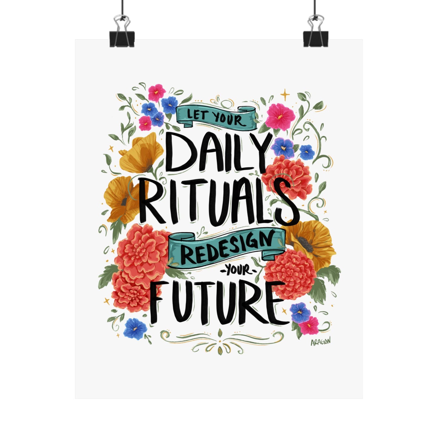 A vertical art print featuring the hand-lettered quote 'LET YOUR DAILY RITUALS REDESIGN YOUR FUTURE' surrounded by colorful watercolor flowers and green vines, shown hanging from two black binder clips against a white background.
