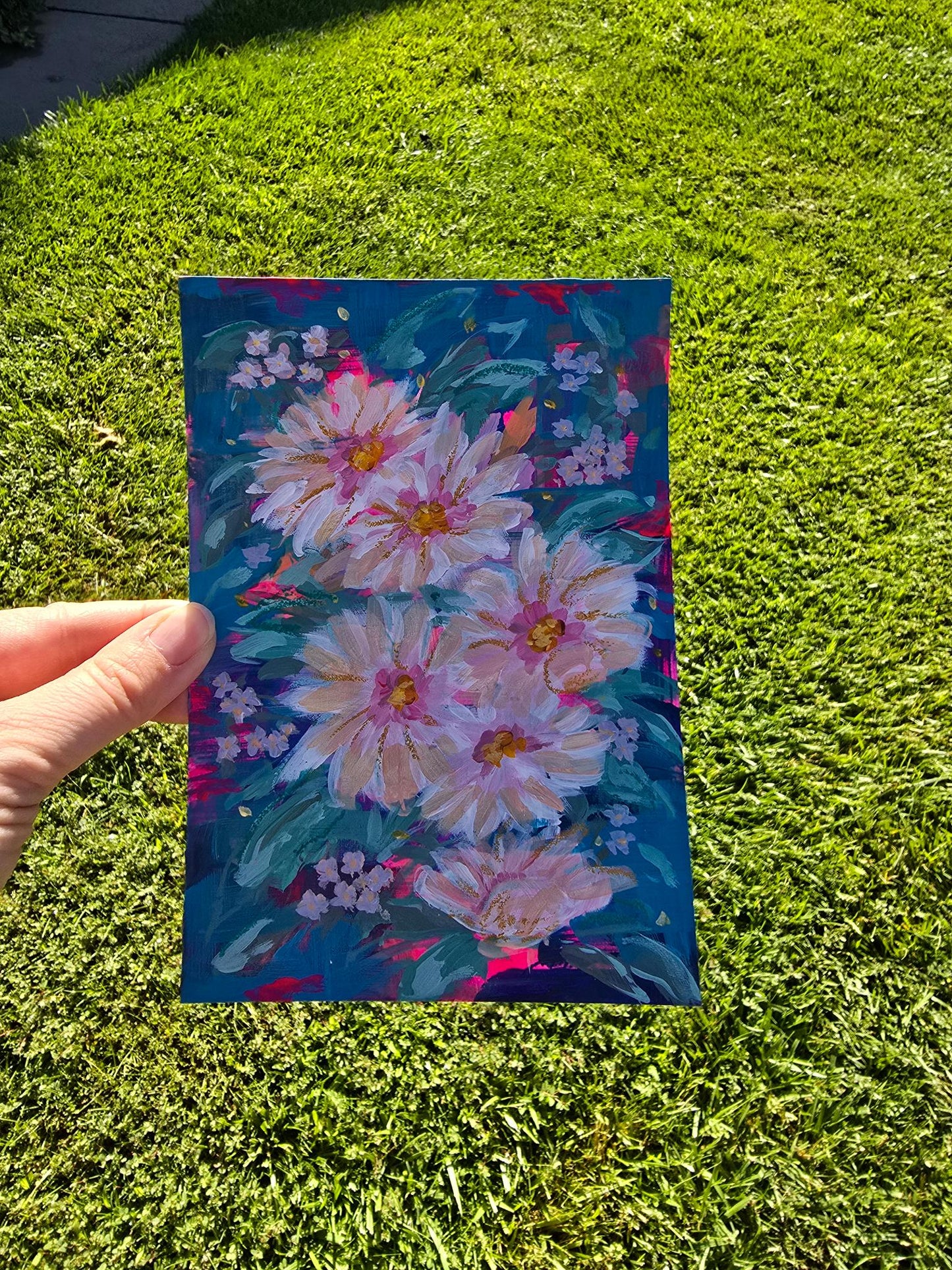 A hand holds a square art print or painting featuring large pink and white flowers with yellow centers on a dark blue background, set against a bright green grass lawn.