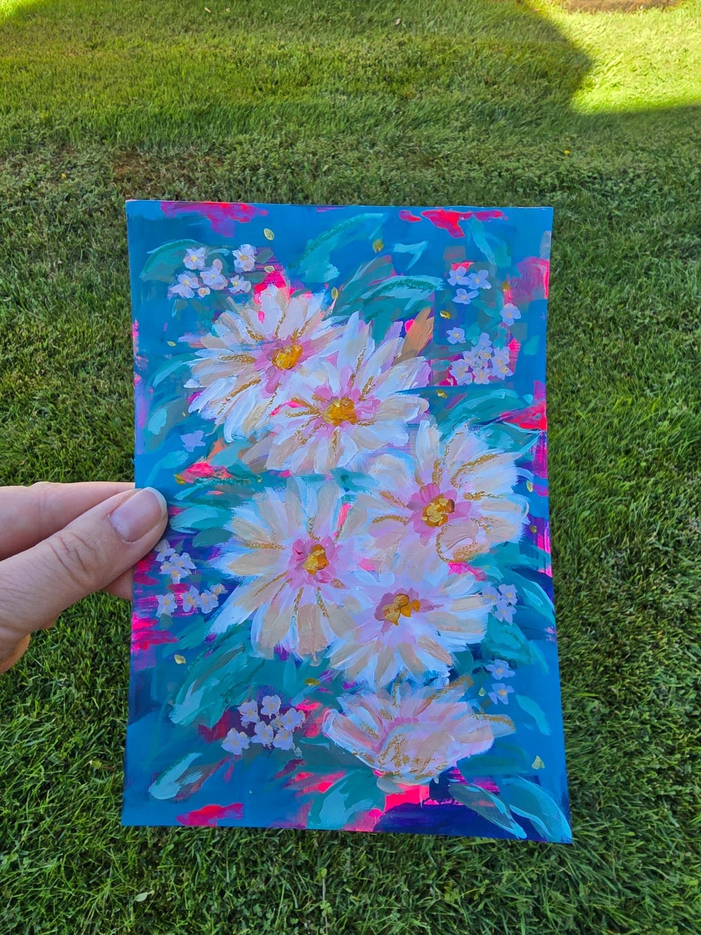 A hand holds a colorful painting of large white and pink flowers with yellow centers and green leaves on a bright blue background, photographed outdoors on a grassy lawn.