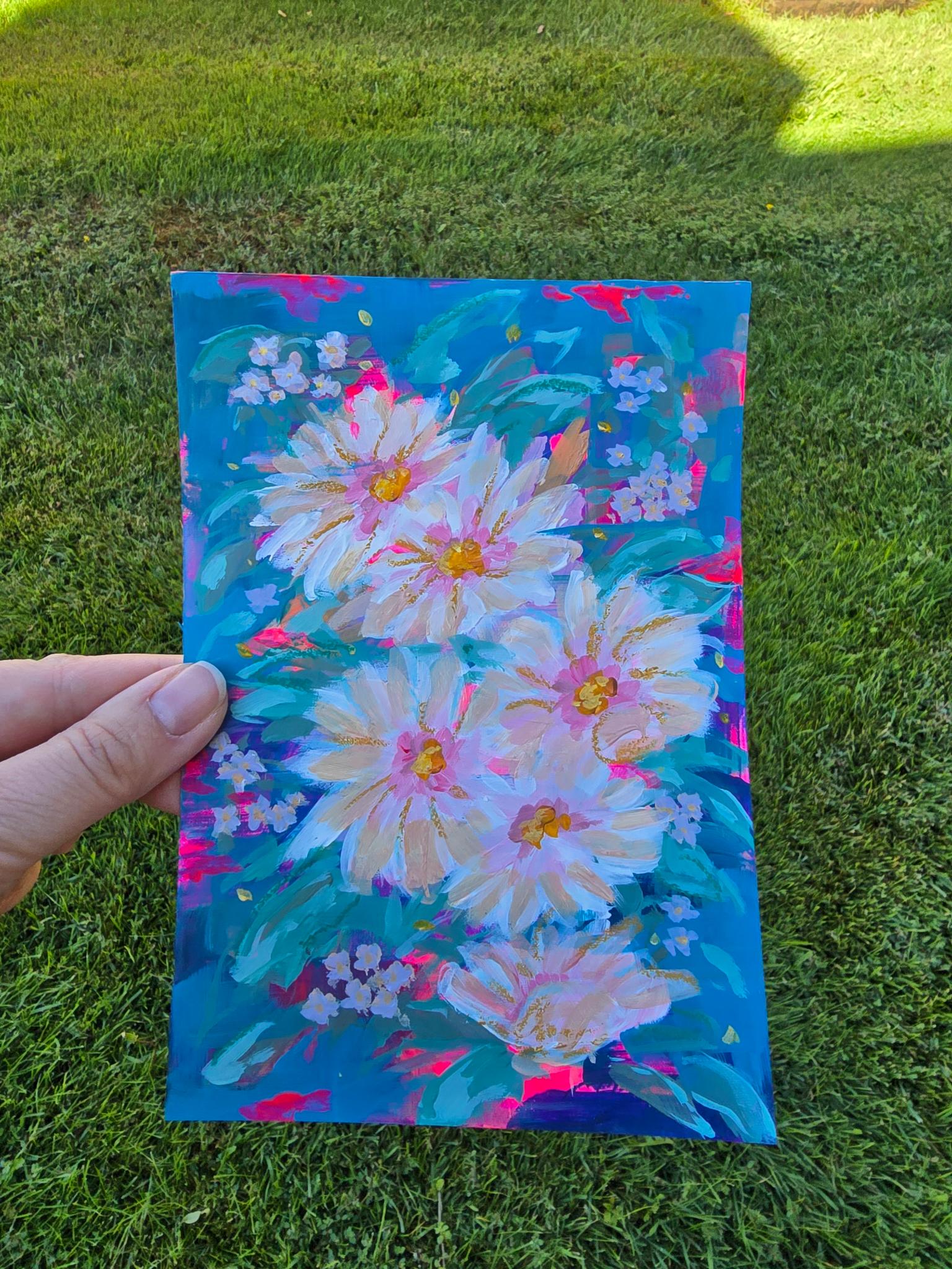 A hand holds a colorful painting of large white and pink flowers with yellow centers and green leaves on a bright blue background, photographed outdoors on a grassy lawn.