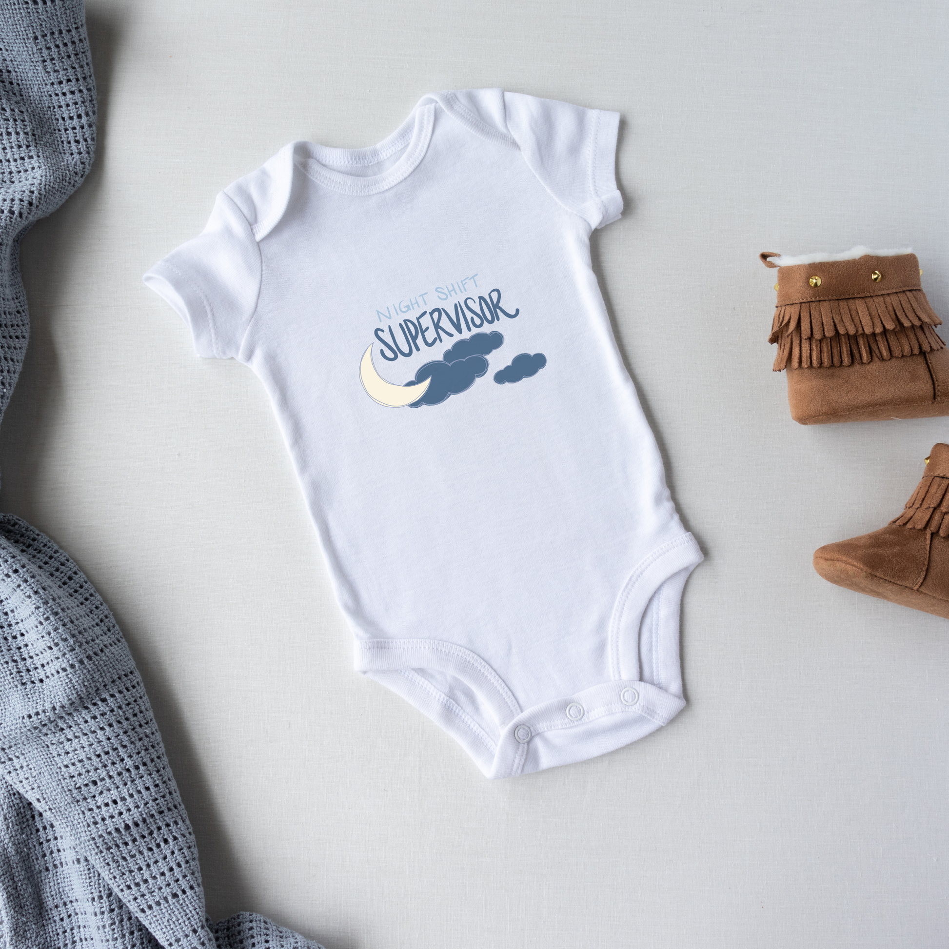 White baby onesie with 'Night Shift Supervisor' text on a light gray surface with a blanket and baby booties.
