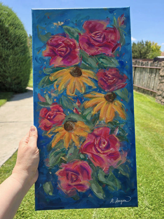 A hand holds a vertical canvas painting outdoors in a garden. The painting features vibrant pink and red roses and yellow black-eyed Susans on a rich blue background.