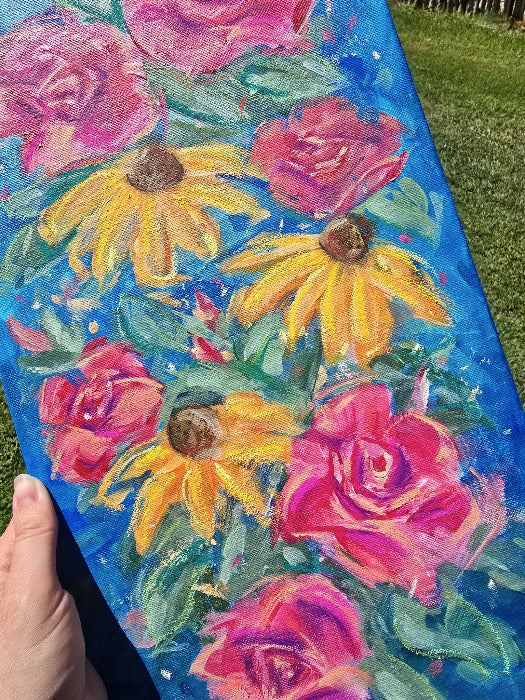 A hand holding a rectangular canvas painting outdoors over green grass. The painting features bright pink roses and yellow coneflowers with green leaves on a vivid blue background.