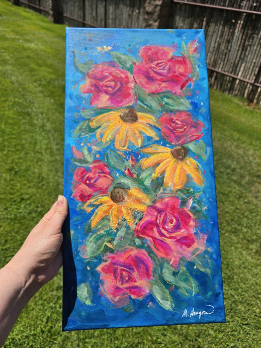 A hand holds a vertical canvas painting of pink roses and yellow flowers on a bright blue background, photographed outdoors on a grassy lawn with a wooden fence behind it.