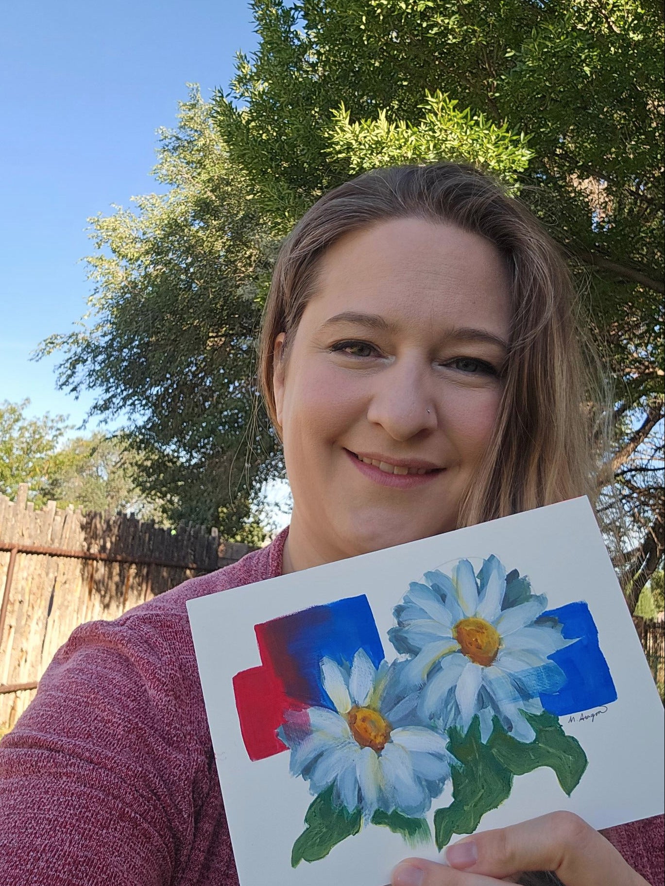 A woman outdoors in a backyard holding up a square painting of two white daisies with yellow centers and green leaves, set against a background of blue sky and green trees.